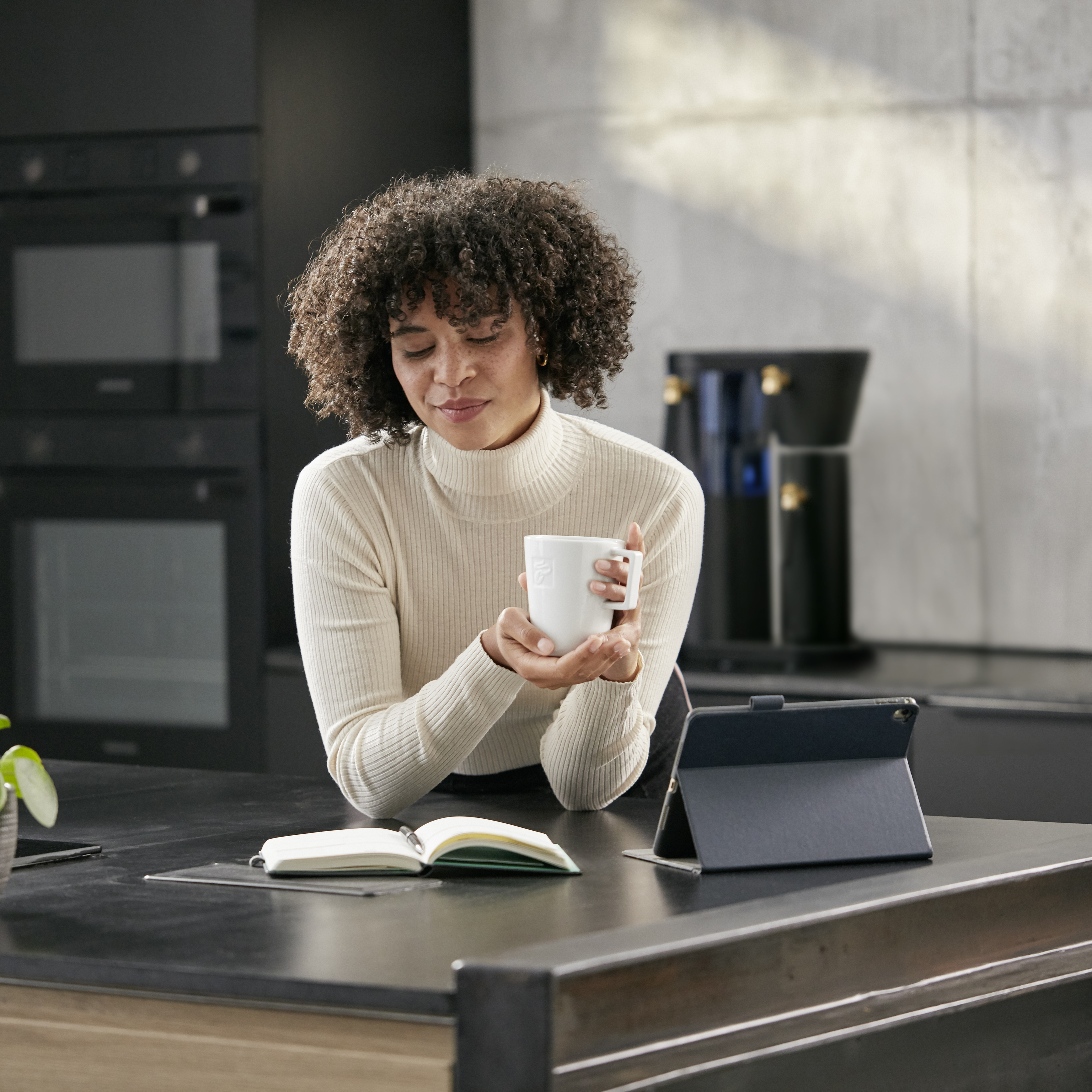 Frau mit Kaffeetasse in einer Küche. Sie liest ein Buch. Im Hintergrund steht eine Filbii. 