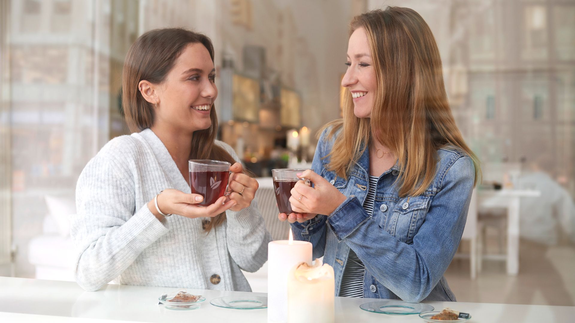 Zwei Frauen hinter einer Glas Wand. Beide schauen sich lächelnd. Beide trinken Tee. 
